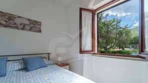 Un dormitorio con cama, almohadas, una mesita de noche, un ventanal con vistas a árboles y una valla.