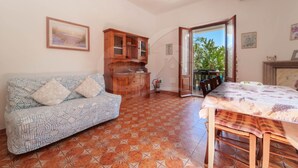 Una habitación con un sofá estampado, un armario de madera, una mesa de comedor con mantel floral y vistas a la vegetación por una puerta abierta.