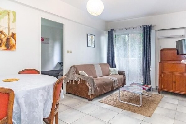 A living room with a brown sofa, a glass coffee table, a dining table with chairs, and a TV on a wooden cabinet. - 1 bedroom apartment in saint-denis (Saint-Denis)