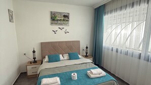 A neatly arranged bedroom with a bed, bedside tables, a hanging light fixture, a wall-mounted artwork, and a window with curtains. - Arslan Homes3 (Muğla)