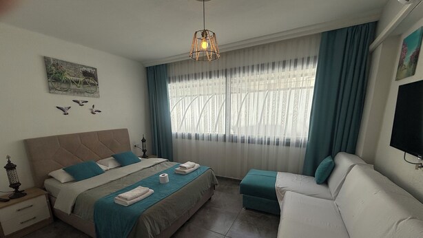 A bedroom with a bed, a sofa, a window with curtains, a wall-mounted TV, and a decorative wall piece. - Arslan Homes3 (Muğla)