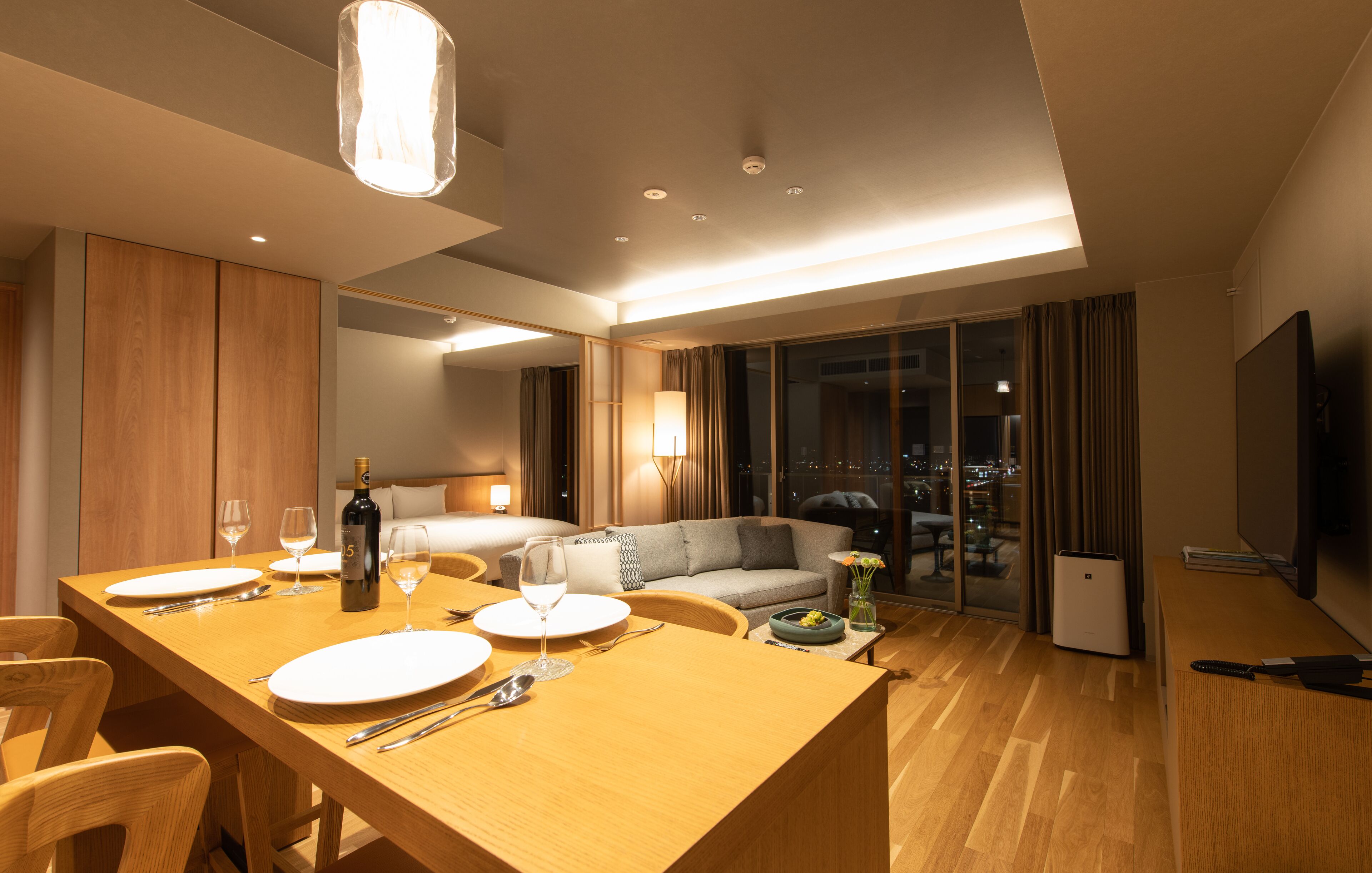 A modern hotel room with a wooden dining table set for two, a sofa, a TV, and a balcony with a city view.