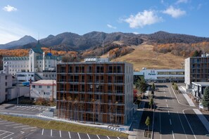 A modern building with a grid-like facade, a parking lot, and a view of mountains in the background. - Fenix West (Furano)