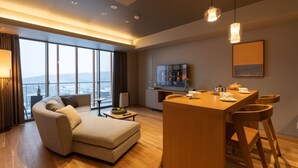 Ein modernes Wohnzimmer mit einer Couch, einem Essbereich mit Holztisch und Stühlen, einem an der Wand montierten Fernseher und großen Fenstern mit Stadtblick.
