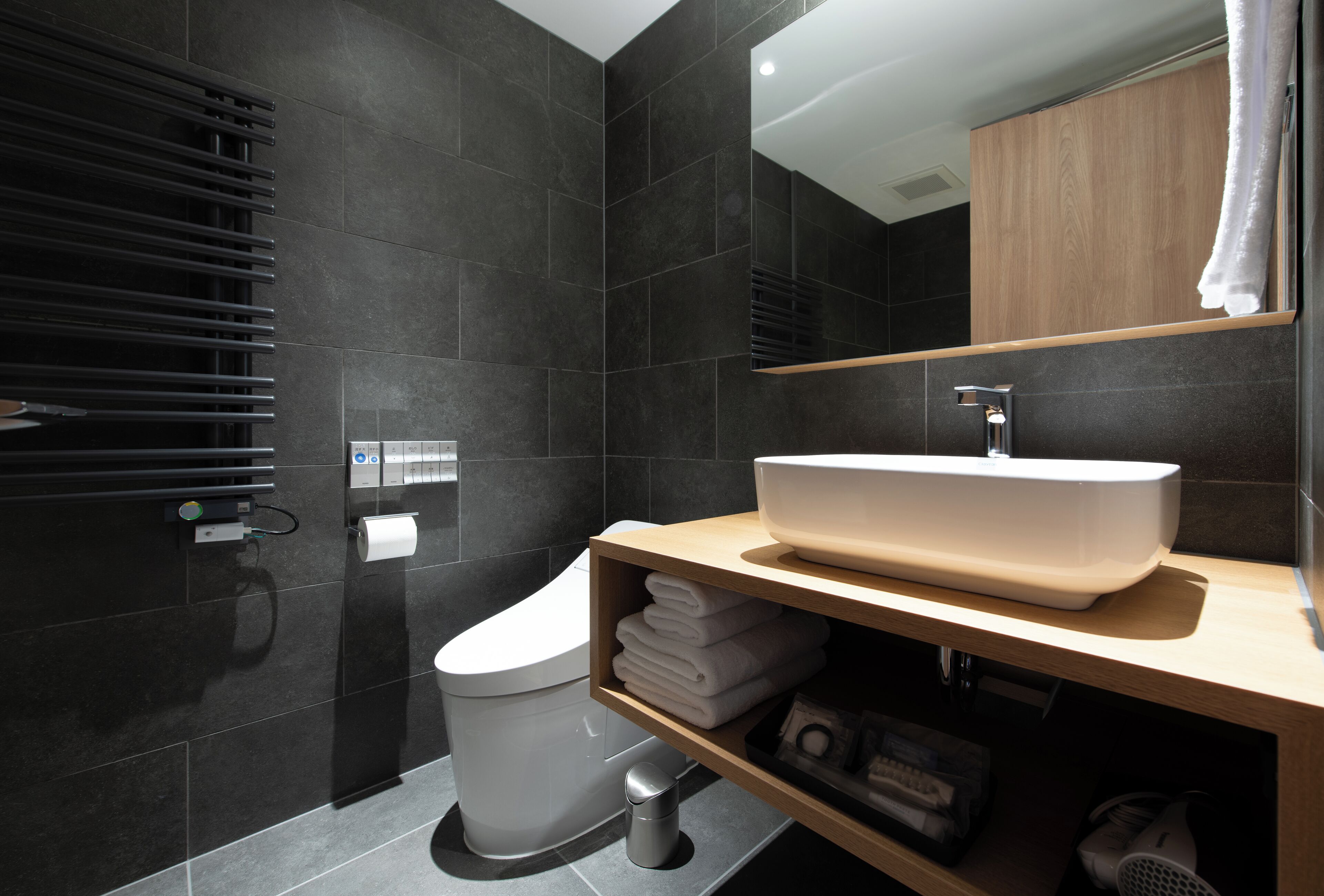 Ein modernes Badezimmer mit dunklen Fliesen, einer weißen Toilette, einem hölzernen Waschtisch mit großem Becken und einem Handtuchhalter.