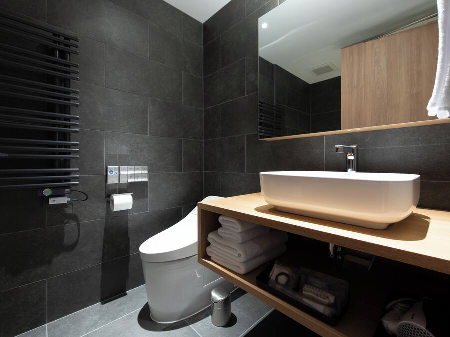 A modern bathroom with dark tiles, a white toilet, a wooden vanity with a large sink, and a towel rack.