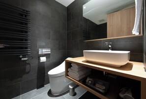 A modern bathroom with dark tiles, a white toilet, a wooden vanity with a large sink, and a towel rack.