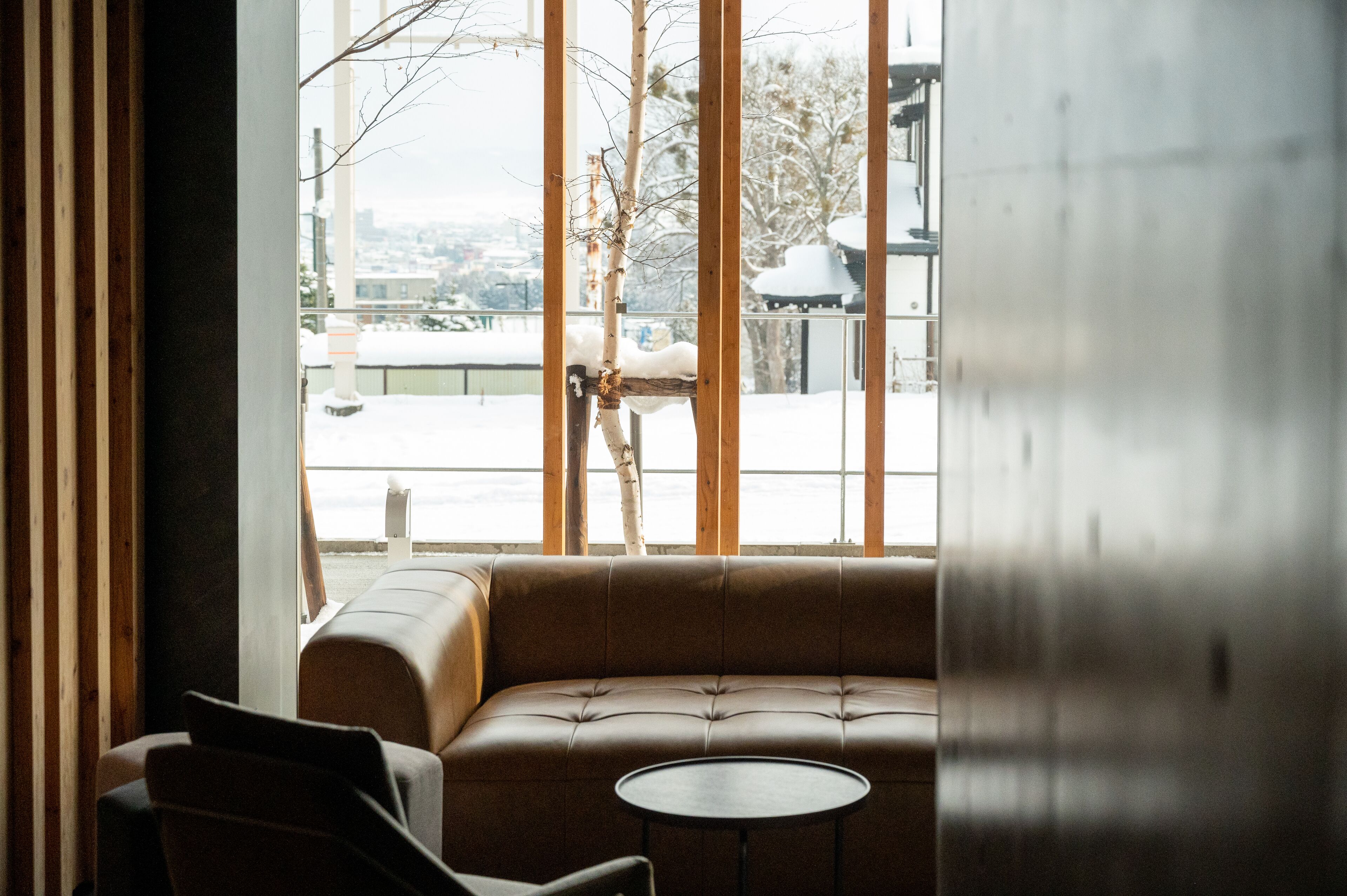 Un interior moderno con un sofá de cuero y una pequeña mesa redonda, ventanas amplias con vistas a un paisaje nevado y edificios.