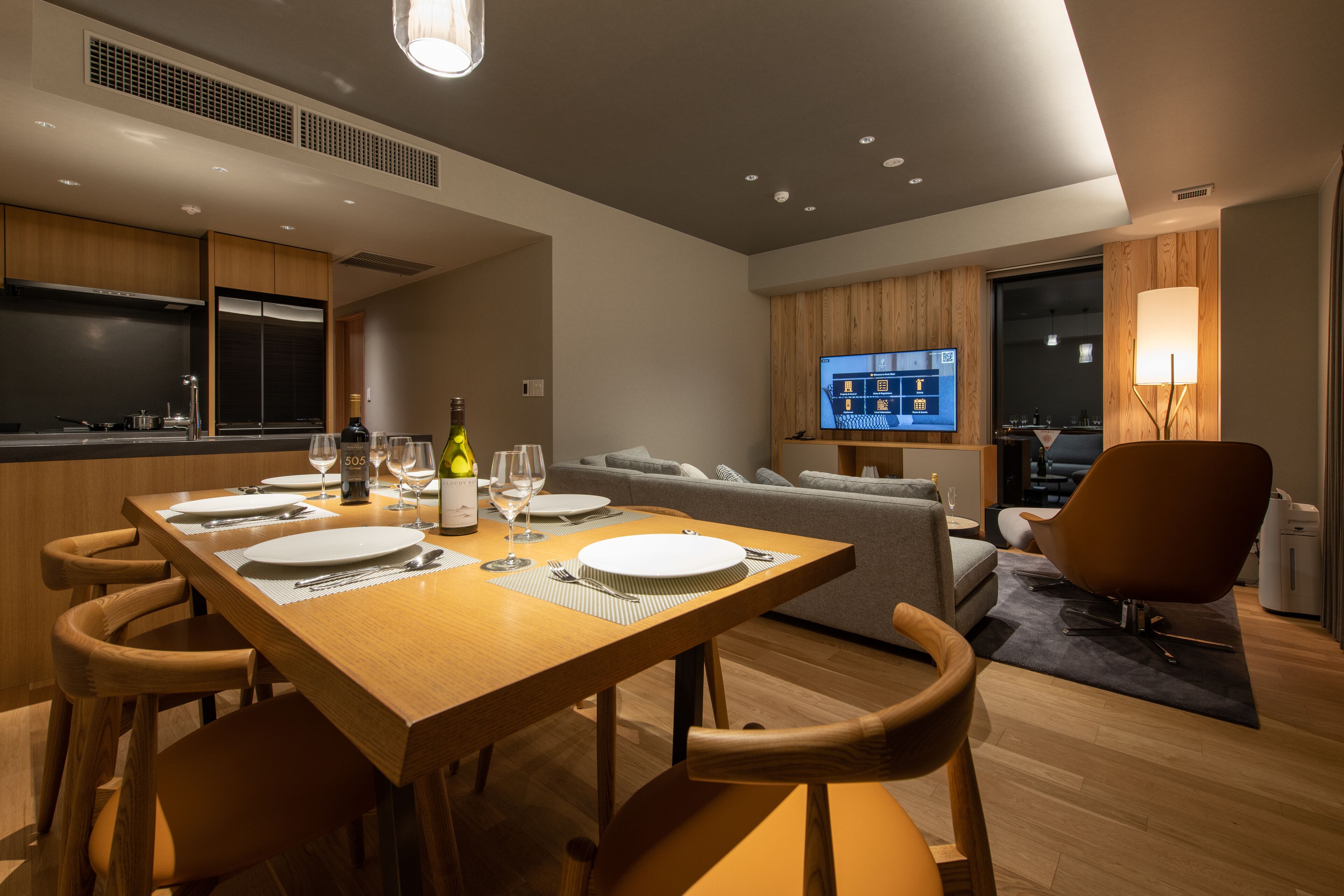 Una sala de estar moderna con una mesa de comedor de madera preparada para cuatro personas, un sofá y un televisor montado en la pared.
