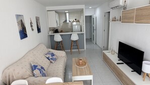 A modern living room with a sofa, coffee table, TV, and a kitchen in the background. - Apartamento Gavines, Miami Playa (Miami Playa)