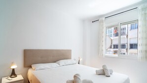 A clean, minimalist bedroom with a large bed, a bedside lamp, and a view of buildings through a window.