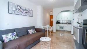 A modern living room with a grey sofa, a small round table, and a kitchen area with white cabinets and a stove.