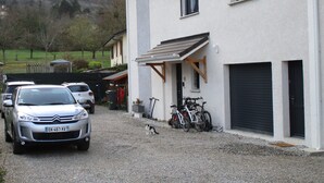 Une allée de gravier mène à un bâtiment blanc doté d’un garage, où sont garés des vélos et des voitures.