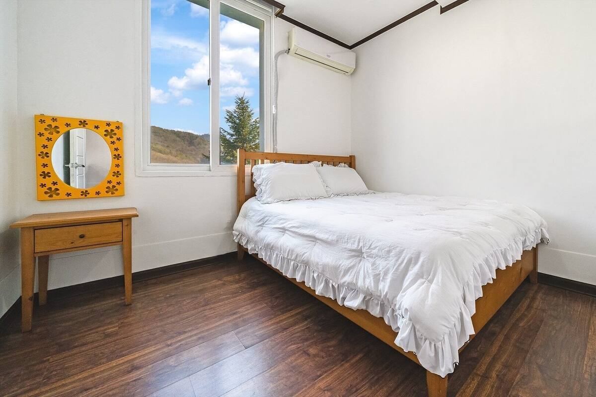 Une chambre avec un lit en bois, une petite table de chevet, un miroir avec un cadre floral et une fenêtre équipée d’un appareil de climatisation.
