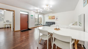 Una habitación de hotel moderna con suelo de madera, una mesa de comedor blanca con sillas, un sofá de cuero negro, una cama con ropa de cama blanca y un ventanal con vistas a árboles.
