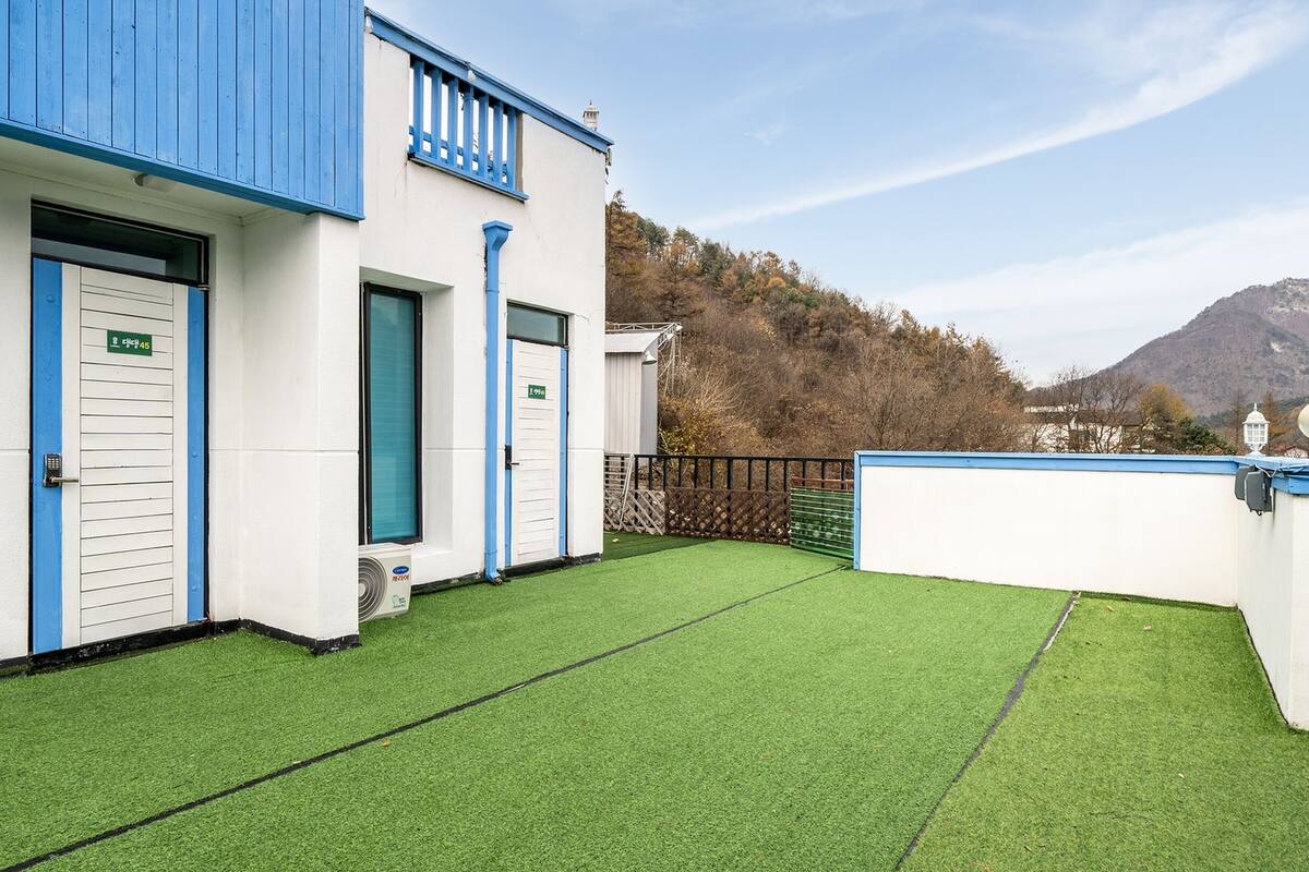 Un bâtiment aux portes bleues et doté d’un balcon, entouré d’une pelouse verdoyante et avec une montagne en arrière-plan.