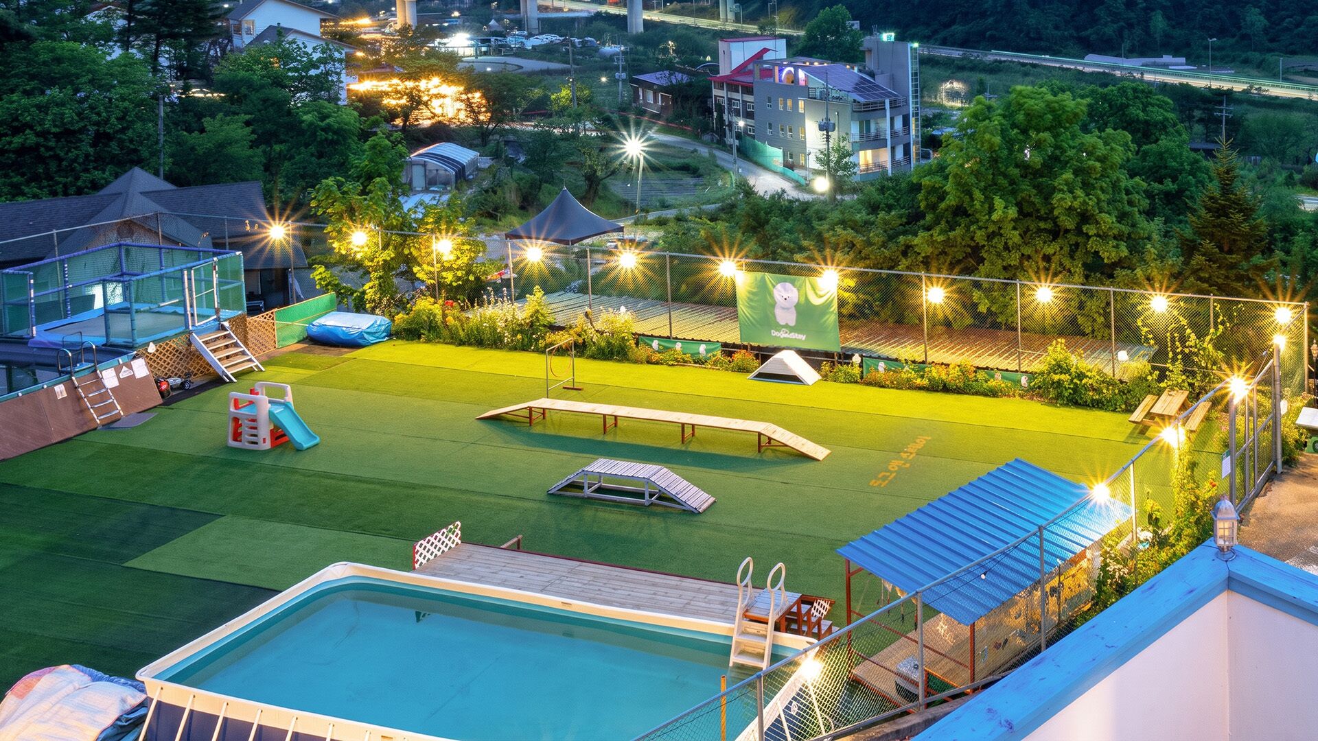 A swimming pool with a slide, a tennis court, and a fenced area with string lights.