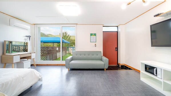 A modern hotel room with a bed, a sofa, a desk with a computer, a TV, and a sliding glass door leading to a balcony.
