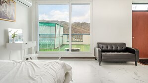 Un dormitorio moderno con un ventanal grande, un sillón de cuero gris, una cama blanca y una mesita de noche con un florero.