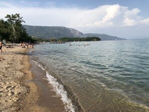 A beach with clear water, people swimming and walking, and mountains in the background.