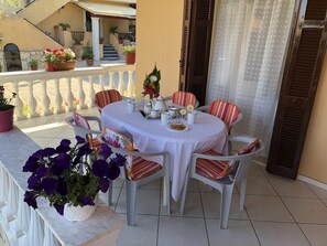 A round table with a white tablecloth, surrounded by striped chairs, set on a balcony with potted plants and a view of a building.