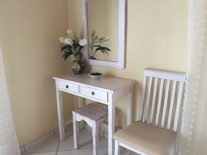 A white desk with two drawers and a small bench, a vase with flowers, and a mirror on the wall.