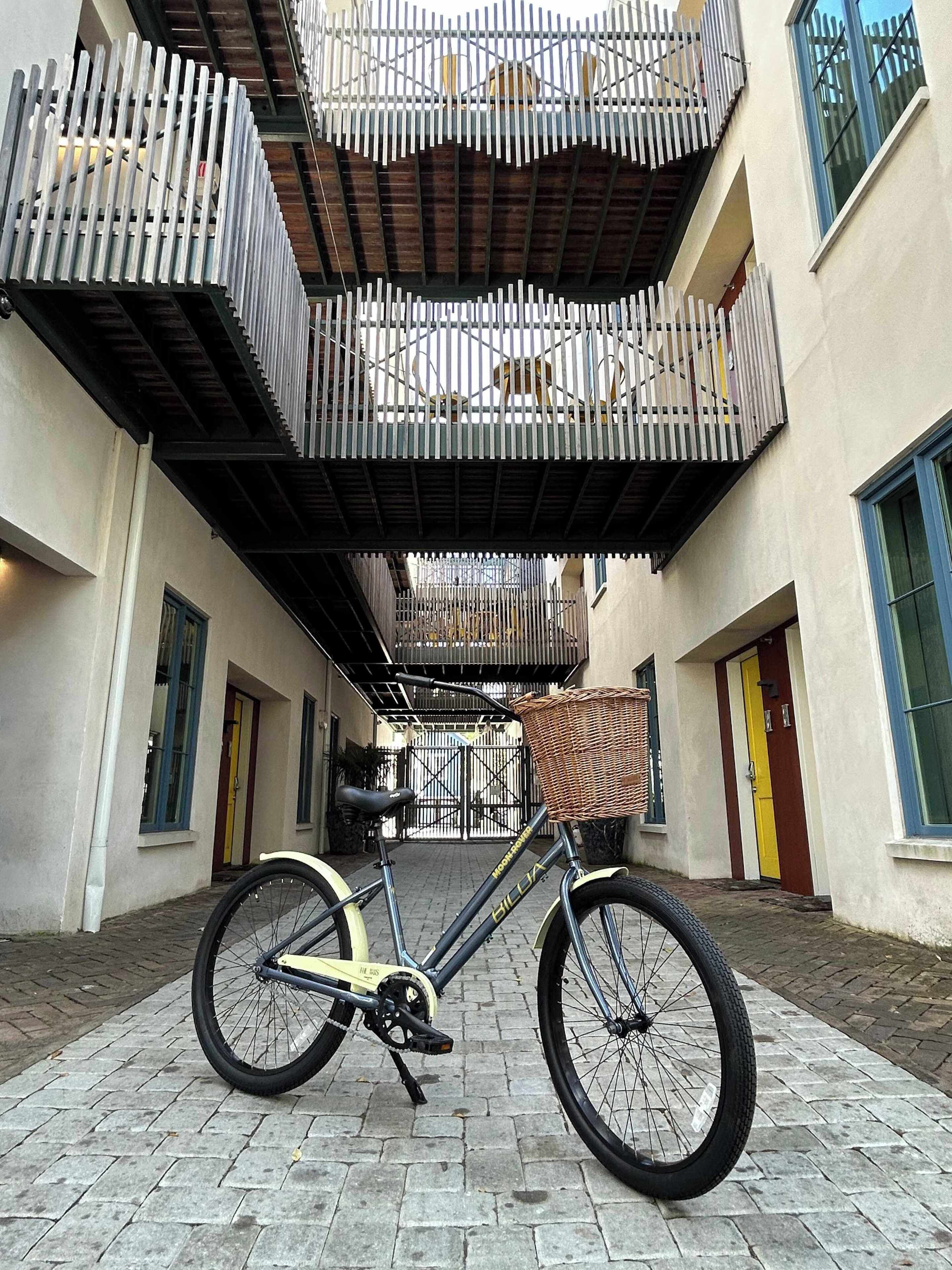 Una bicicleta con cesto estacionada en un sendero adoquinado frente a un edificio con entrada protegida y un balcón con barandilla.