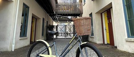 Una bicicleta con cesto estacionada en un sendero adoquinado frente a un edificio con entrada protegida y un balcón con barandilla.