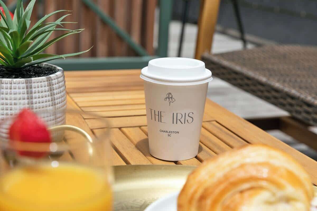 A coffee cup with 'THE IRIS' branding on a wooden table, a potted plant, and a croissant.