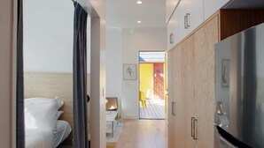 Un couloir étroit, avec un lit sur la gauche, une petite cuisine sur la droite et une porte donnant sur une pièce lumineuse.