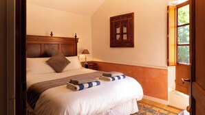 A neatly made bed with pillows and a folded blanket, a wooden headboard, a bedside table with a lamp, and a window with a view of greenery.