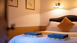 Una cama bien hecha con un cojín blanco y amarillo, una colcha a rayas azules y amarillas, y una toalla azul doblada. Hay una lámpara de pared sobre la cama.