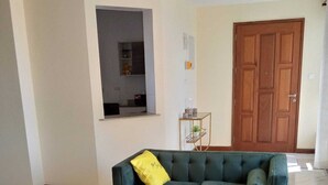 Sala de estar con un sofá verde, una mesa de centro de vidrio, una puerta de madera y una mesita con objetos decorativos.