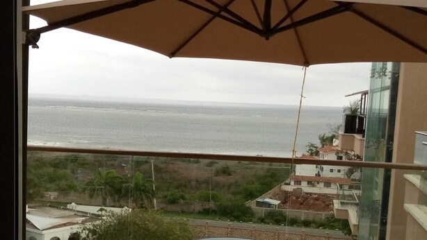 Un balcón con una mesa y dos sillas, con vista al mar y edificios, rodeado de vegetación.