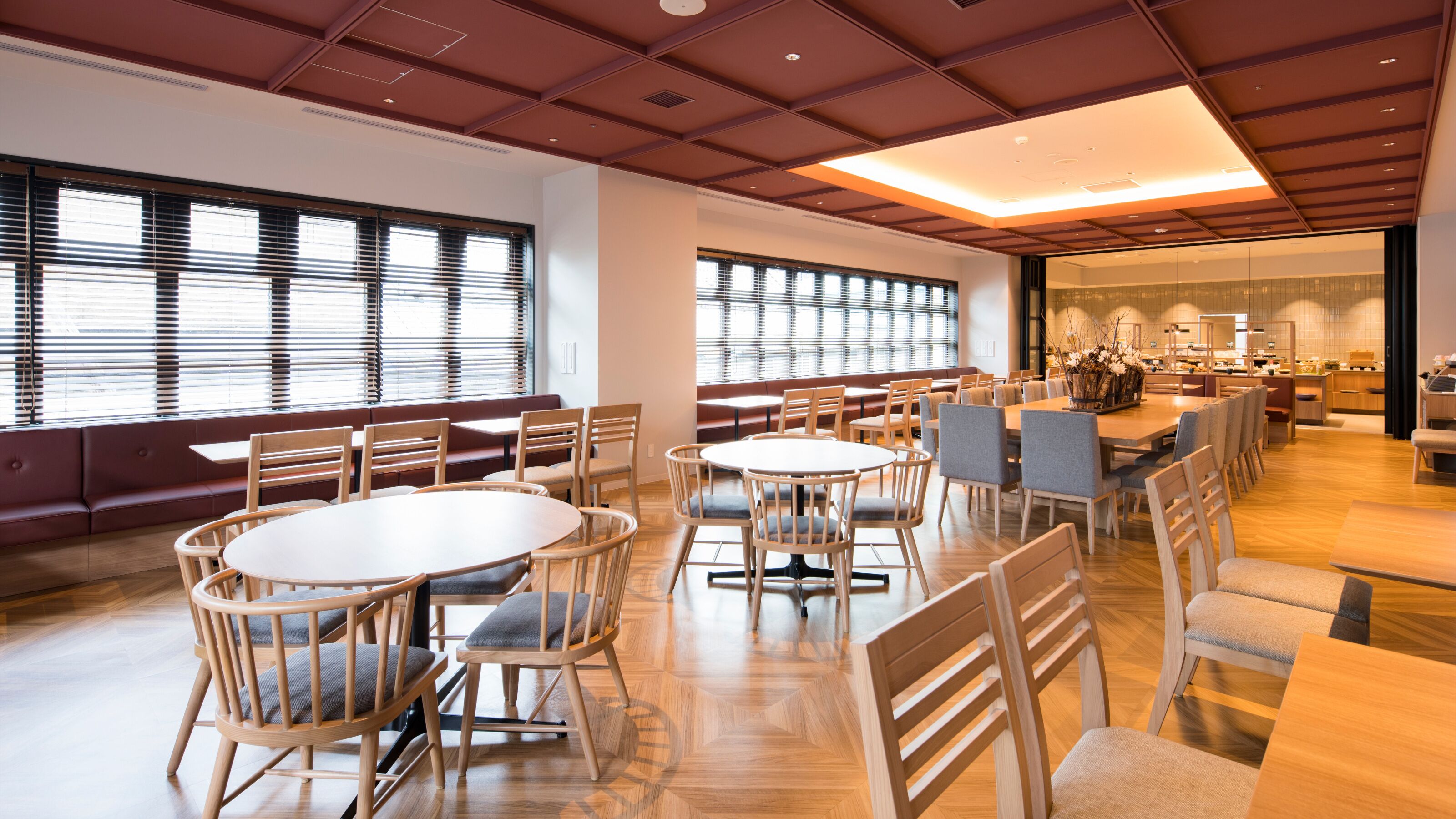 A spacious dining area with multiple round tables and chairs, large windows with blinds, and a modern ceiling design.