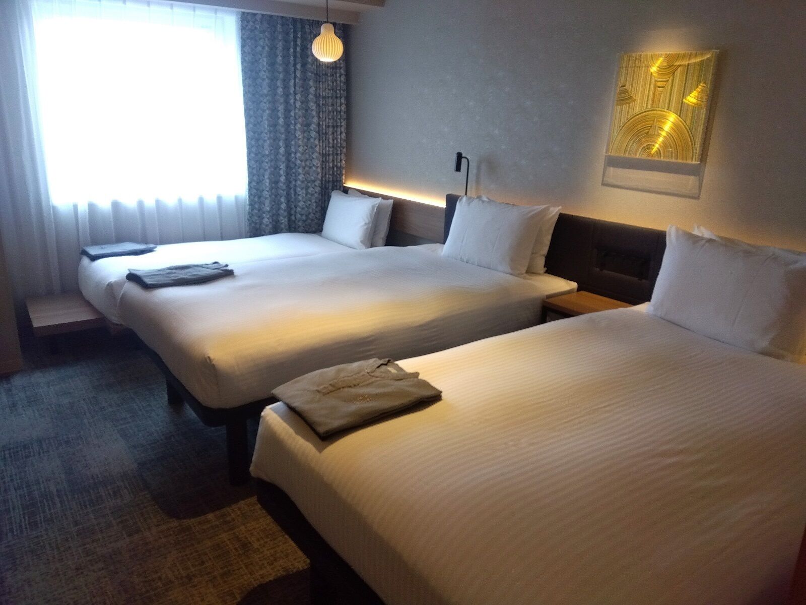 Hotellihuone, jossa on kaksi sänkyä, työpöytä, tuoli, lamppu ja ikkuna, jossa on verhot.