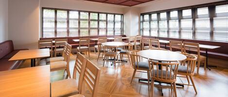 Uma sala de jantar ampla, com várias mesas redondas e cadeiras de madeira, grandes janelas com persianas e piso de madeira.