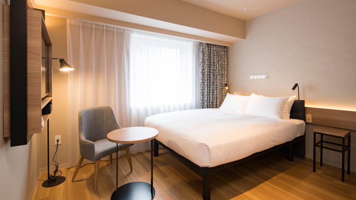 Et hotelværelse med en seng, en stol, et lille bord, et skrivebord og et fjernsyn.