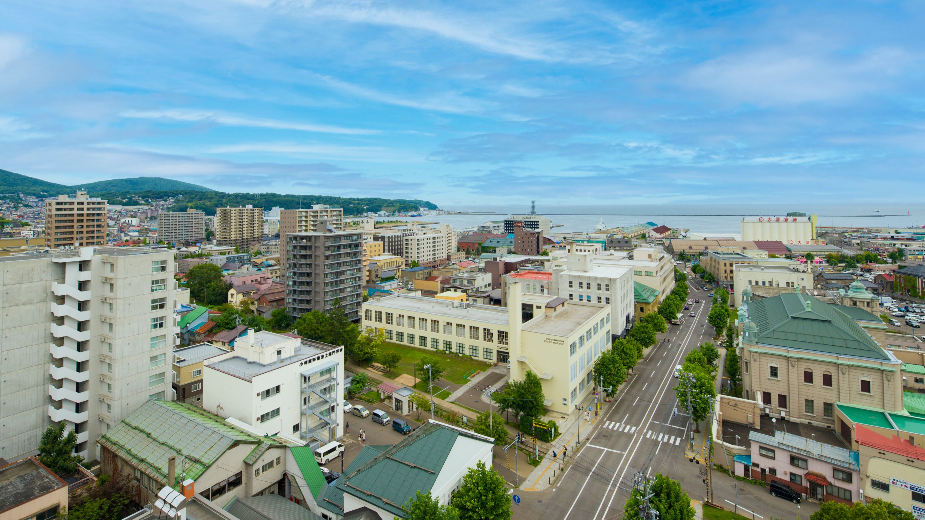A cityscape with a mix of modern and older buildings, a wide street, and a view of the sea in the distance.