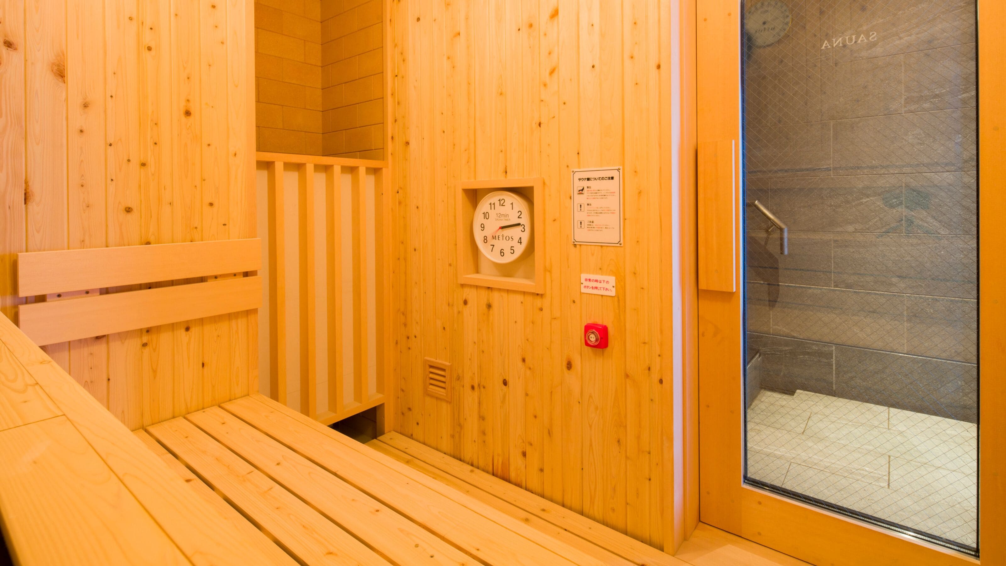 A wooden sauna room with a bench, a clock, and a glass door leading to a tiled area.