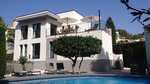 Un edificio moderno de dos pisos con piscina, mobiliario exterior y un gran árbol.
