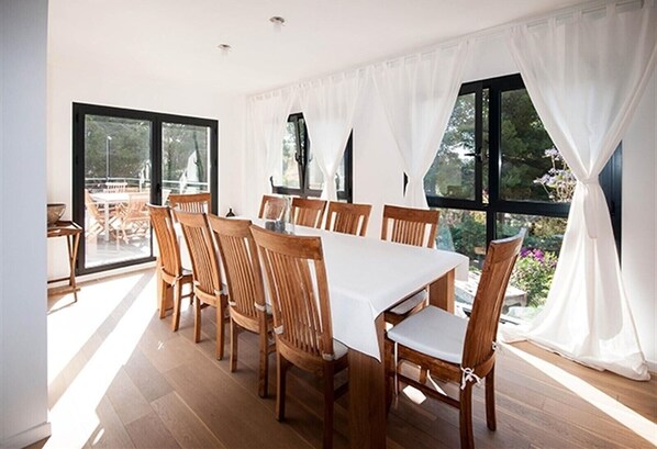 A dining area with a large table and wooden chairs, large windows with white curtains, and a view of outdoor furniture.