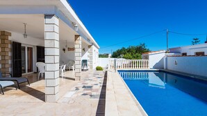Un área junto a la piscina con cielo azul despejado, un edificio blanco con patio de baldosas y una pileta.