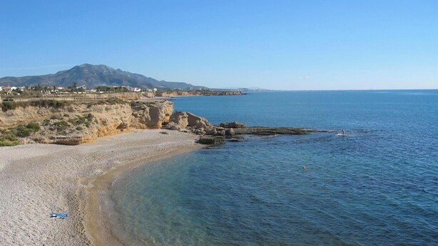 Una playa costera con aguas cristalinas, costa rocosa y una montaña distante.