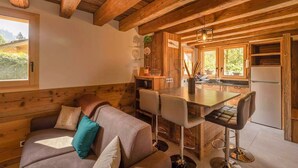 A cozy cabin interior with a wooden ceiling, a dining area with bar stools, a kitchen with a refrigerator, and a sofa with cushions.