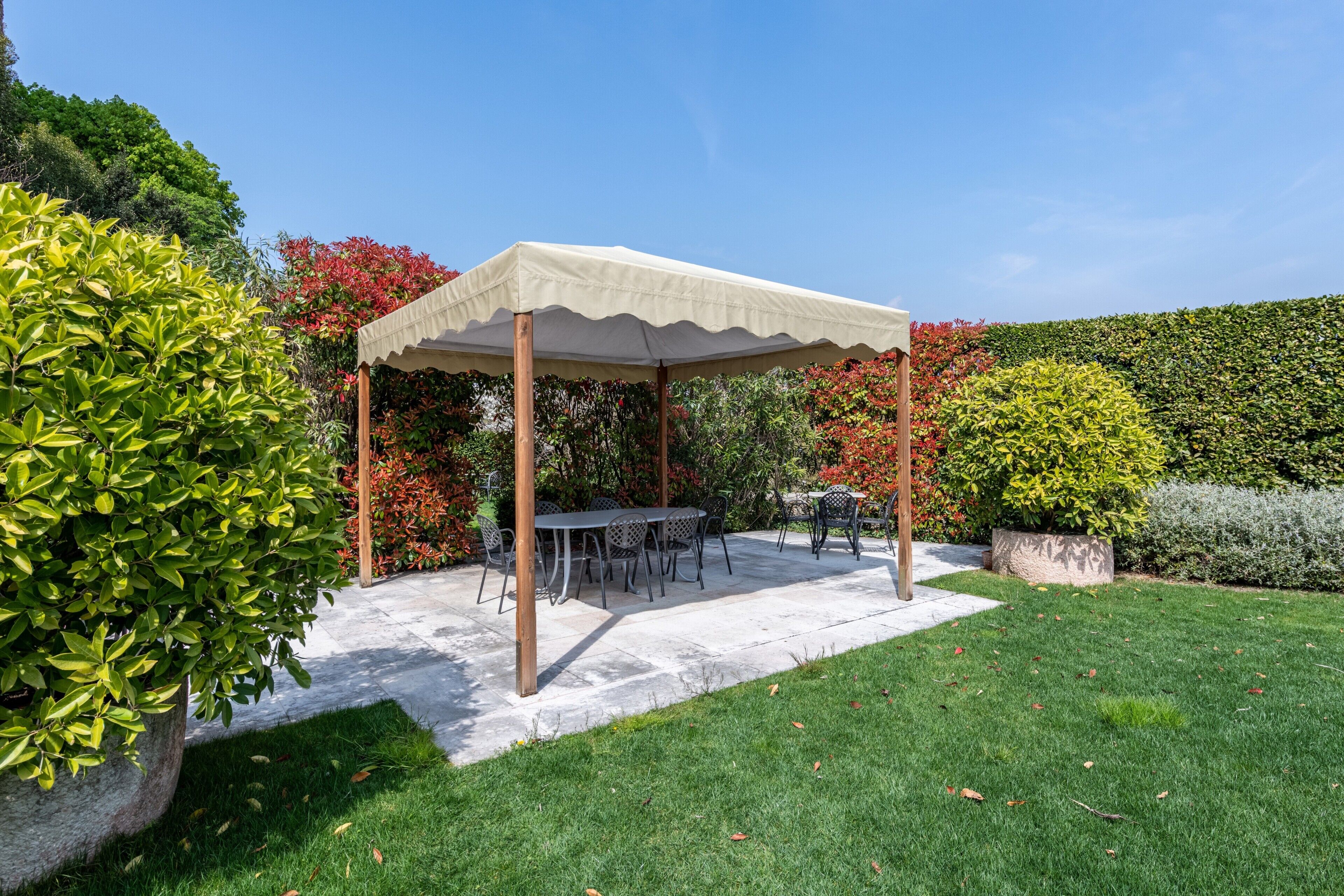 Ein überdachter Patio-Bereich mit Sitzgelegenheiten im Freien, umgeben von gepflegten Hecken und einem Rasen.