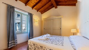 Une chambre avec un plafond en bois, un lit recouvert d’une couvre-lit à motifs floraux, une fenêtre avec des rideaux et une lampe fixée au mur.