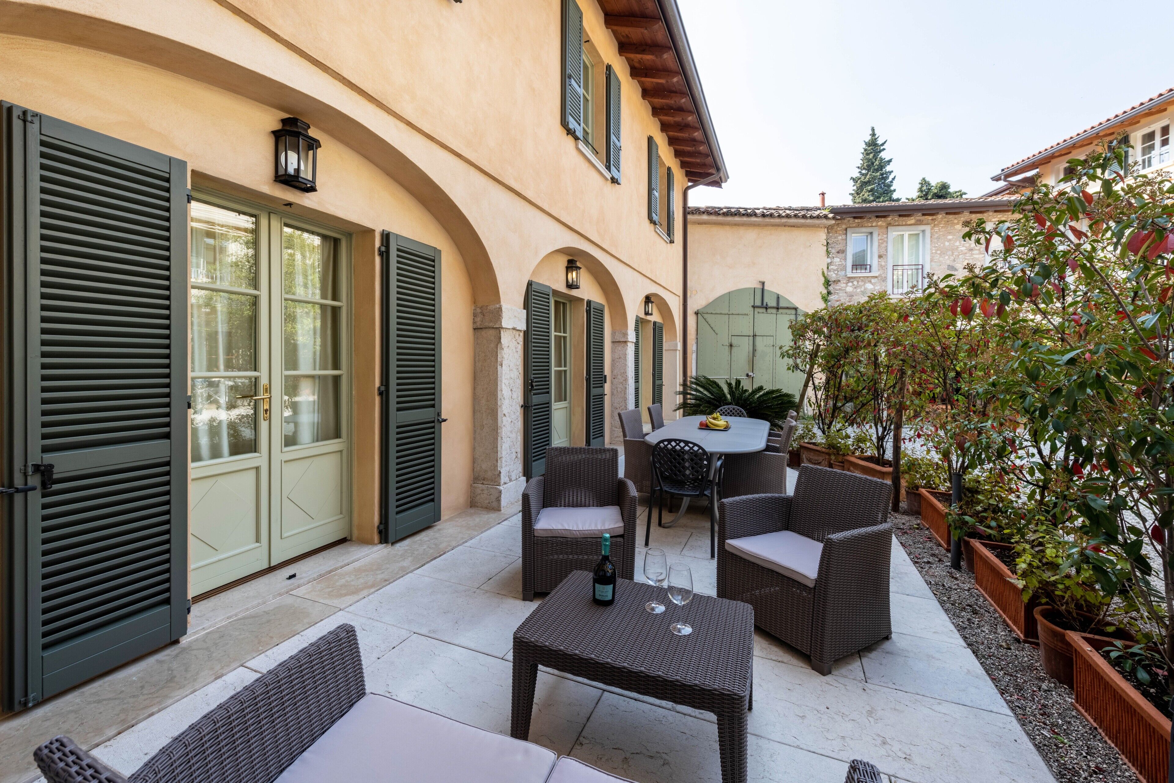 Une terrasse extérieure avec des meubles en osier, une table avec une bouteille et un verre, et une vue sur un bâtiment doté d’arcades et de volets.