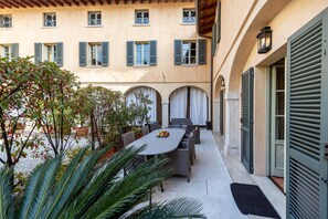 Un patio al aire libre con una mesa y sillas, rodeado de plantas en macetas y un edificio con ventanas arqueadas y contraventanas.