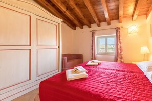 A bedroom with a red bedspread, a wooden ceiling, a window with curtains, and a bedside lamp. - Apartment 'Antica Torre Goethe' with Mountain View, Wi-Fi and Air Conditioning (San Felice del Benaco (BS))